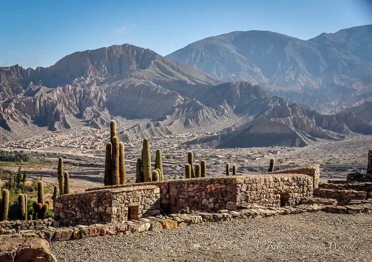 A stone brick wall has cacti behind it, and behind that are a series of jagged mountains. This is Tilcara in Northwest Argentina, a great stop on a 14 day Argentina trip