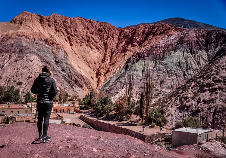 A woman in black stands on a hill overlooking the Cerro de Siete Colores - a hill with multi-coloured layers. It's one of the most popular places to visit in Northwest Argentina