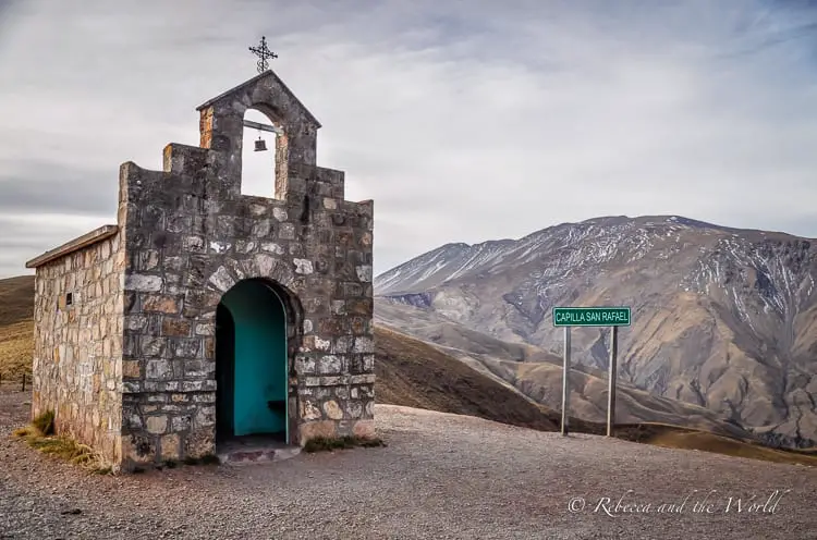 A small stone church sits on a hill. It. has a green door and a green road sign next to it says Capilla San Rafael. Behind the church are mountains. This small chapel is in northwest Argentina - a great addition to two weeks in Argentina