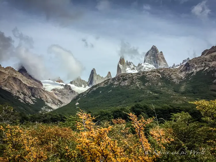 A view of Mount Fitz Roy shows yellow leaves in front of the rocky peaks, with lush green trees in between.