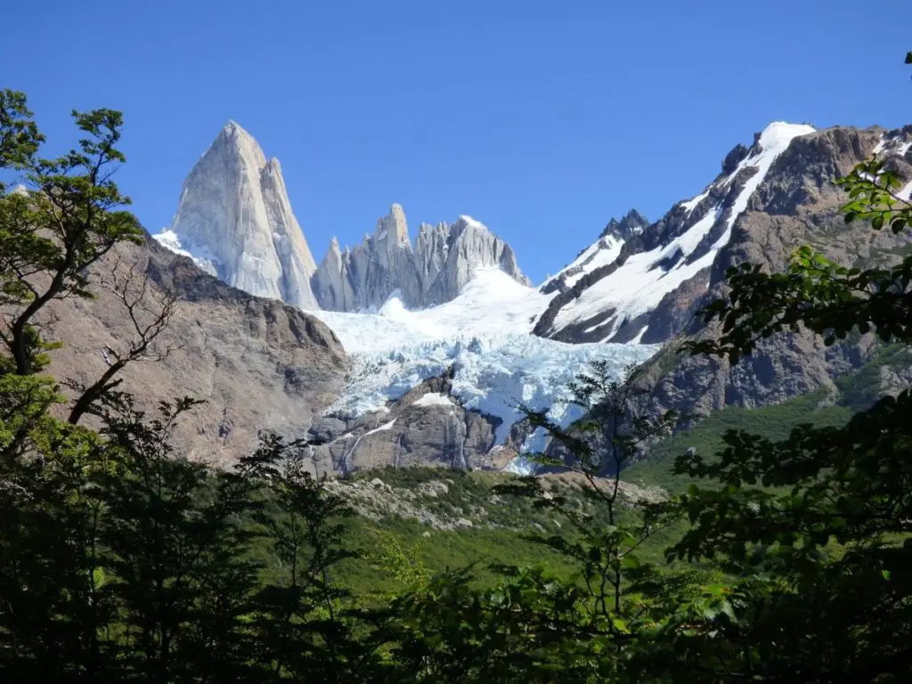 Mount Fitz Roy - with snow and glaciers through the rocky peaks - is framed through green trees. El Chalten is a great honeymoon destination in Argentina for couples seeking adventure and hiking.