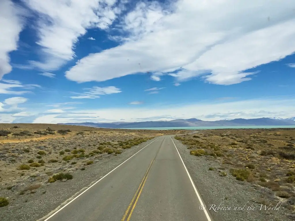 An open road stretches through the arid Patagonian landscape under a vast sky with dynamic cloud formations, leading towards distant mountains and a lake. The journey from El Calafate to El Chalten is really beautiful.