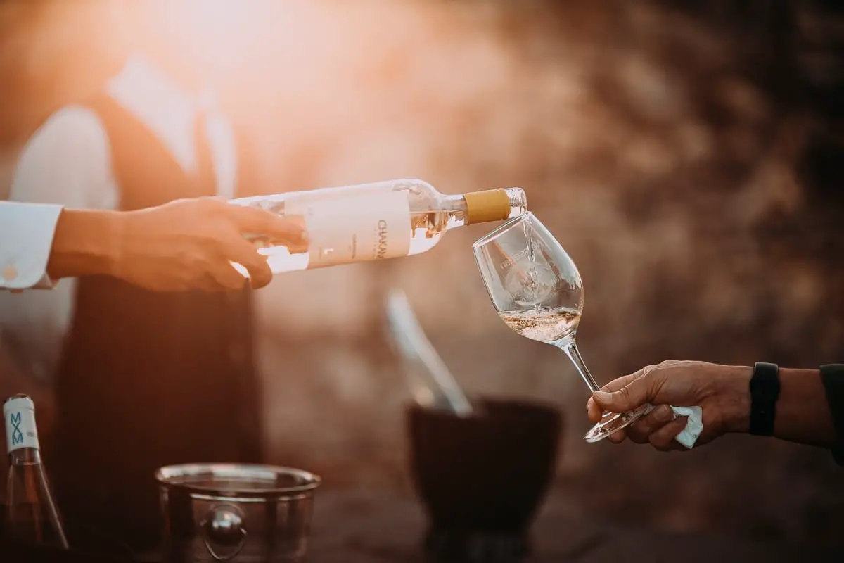 A person pouring white wine from a bottle into a wine glass held by another person, with a blurred background suggesting an outdoor setting. There's always plenty of wine served at estancias in Argentina, especially the luxury and high-end estancias.