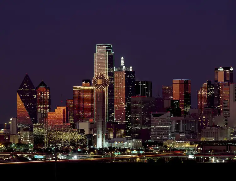 The Dallas skyline at night, featuring a variety of illuminated high-rise buildings. The tallest building has a green outline, and the prominent Reunion Tower with its spherical top is brightly lit.