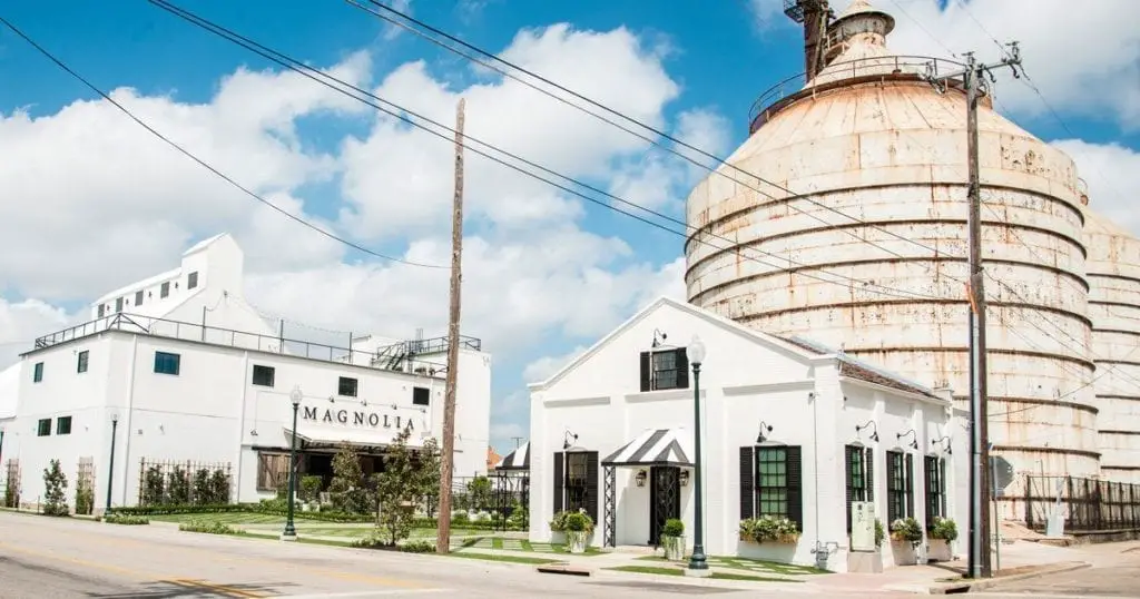 Magnolia Market at the Silos in Waco. The scene includes a white building with a black and white striped awning, large rusty silos, and a neatly landscaped area. The sky is blue with a few clouds, and the street in front of the market is empty.
