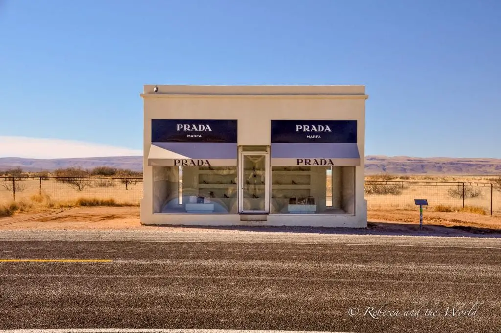 The Prada Marfa art installation, a replica of a Prada storefront, situated in a desert landscape under a clear blue sky. The minimalist building features large display windows with Prada products and black awnings with the Prada logo. One of the quirkiest places to visit in Texas is Marfa.