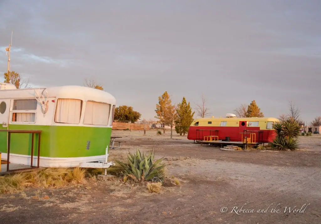 Two vintage trailers in a desert landscape at sunset. One trailer is green and white, while the other is red and yellow. The ground is dry and grassy, with a few scattered plants and trees in the background. El Cosmico is one of the best places to stay in Marfa, Texas.