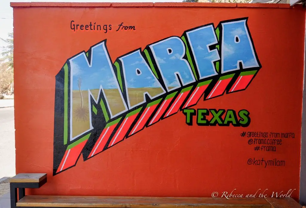 A colorful mural in Marfa, Texas, that reads "Greetings from Marfa" on a bright orange wall. The word "MARFA" is painted in large, bold letters with desert landscape scenes depicted inside the letters. The mural includes hashtags and social media handles at the bottom. Marfa is a unique place to go in Texas.