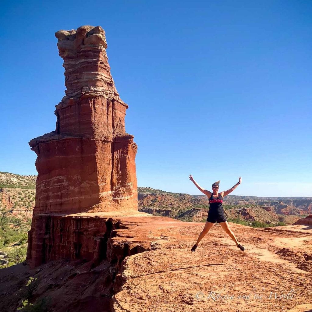A person - the author of this article - jumping in front of the Lighthouse rock formation in Palo Duro Canyon. The rock is a tall, red sandstone pillar with layered bands, set against a clear blue sky. The person is mid-jump with arms and legs outstretched, wearing a black tank top and shorts. Palo Duro Canyon in Texas is a great place for hiking.
