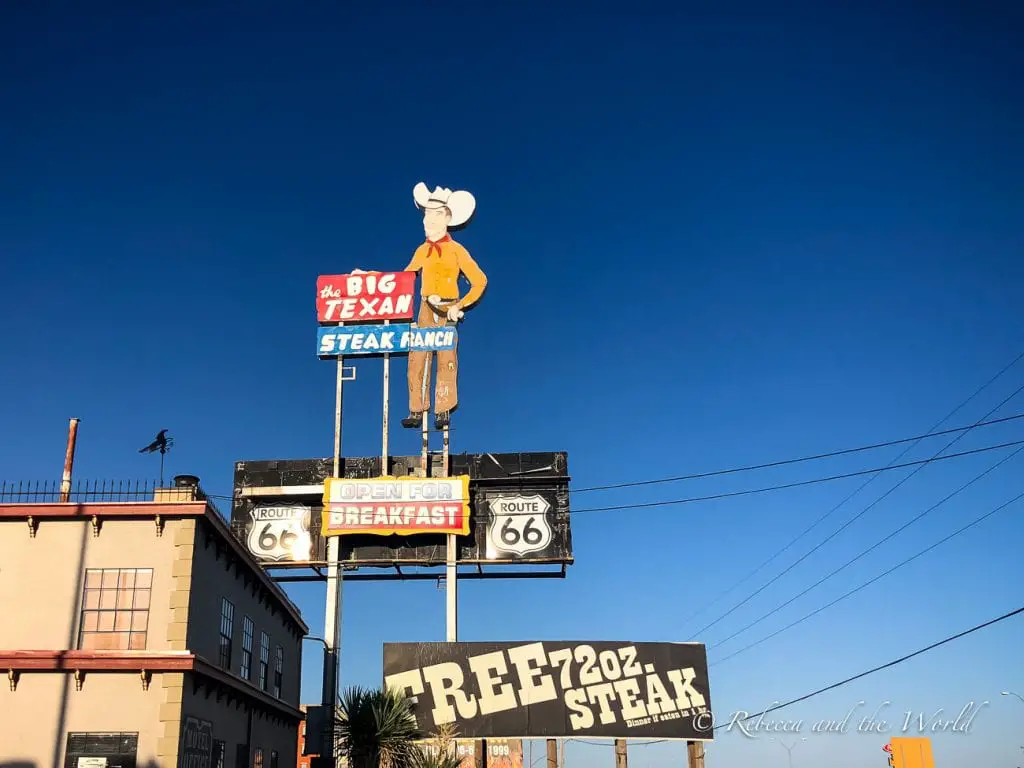 The Big Texan Steak Ranch sign in Amarillo. The sign features a large, colorful cowboy figure and text advertising a "FREE 72oz. STEAK." The sky is clear and blue, with the building partially visible below the sign. The Big Texan in Amarillo, Texas, is famous for its steak-eating challenge.