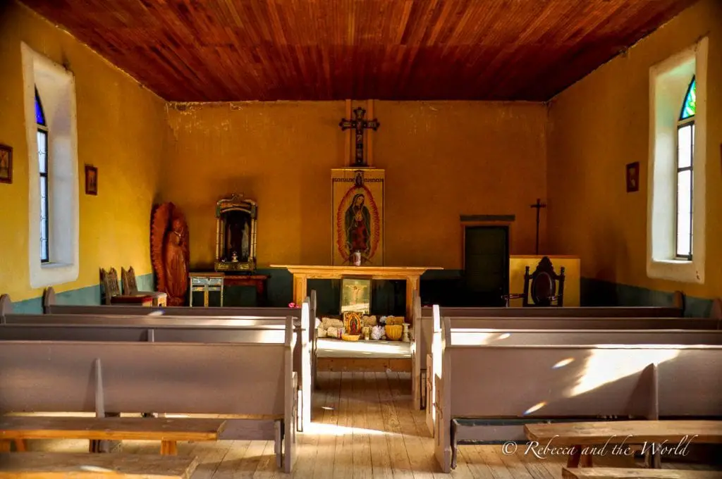 The interior of a small, rustic chapel with wooden pews, a simple altar, and religious decorations. The room has a warm, inviting atmosphere with light streaming through the windows. Terlingua Ghost Town is a quirky place to visit in Texas.