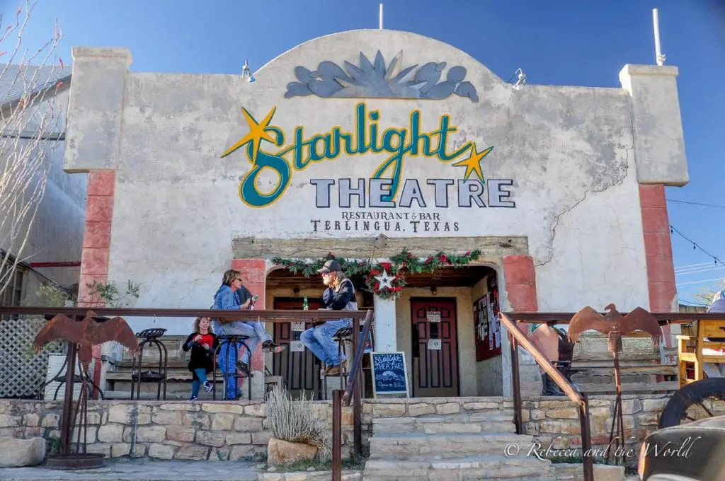 The Starlight Theatre Restaurant & Bar in Terlingua, Texas. The exterior of the building is rustic with a large sign reading "Starlight Theatre" and a few people sitting on the front steps under a clear blue sky. The Starlight Theatre in Terlingua, Texas, is a great place for music and good food.