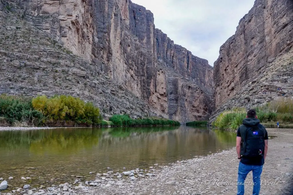 A scenic view of the Santa Elena Canyon in Big Bend National Park. The photo shows tall, rugged cliffs surrounding a calm river, with greenery along the riverbanks and a person - the author's husband - standing in the foreground looking at the canyon. Big Bend National Park has some of the most beautiful views in Texas.