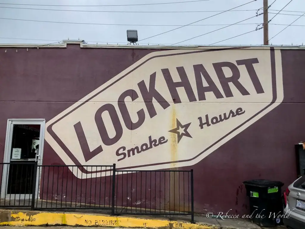A wall painted with the words "Lockhart Smokehouse" in large, bold lettering. The wall is purple, and the text is enclosed in a shape resembling a vintage sign. A door and railing are visible on the left side. You can't visit Texas without trying barbecue.