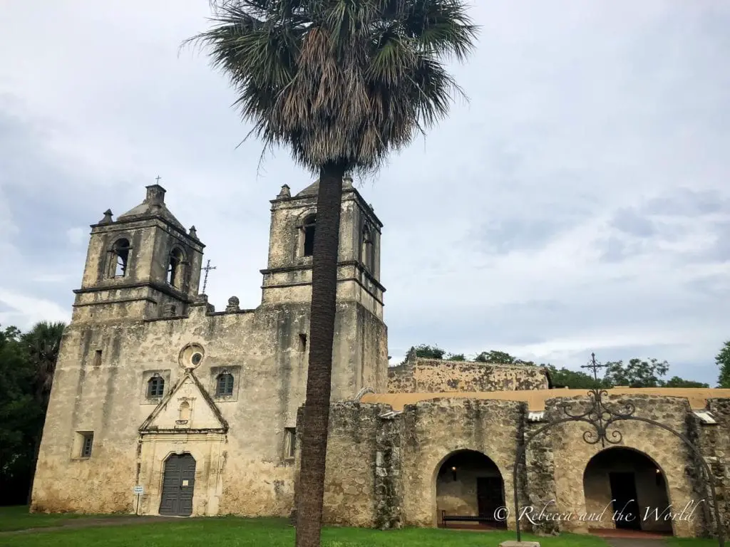 The historic Mission Concepción in San Antonio, an old stone church with twin bell towers and a palm tree in the foreground. The sky is overcast, adding to the serene atmosphere of the site. San Antonio has several historic missions to visit.
