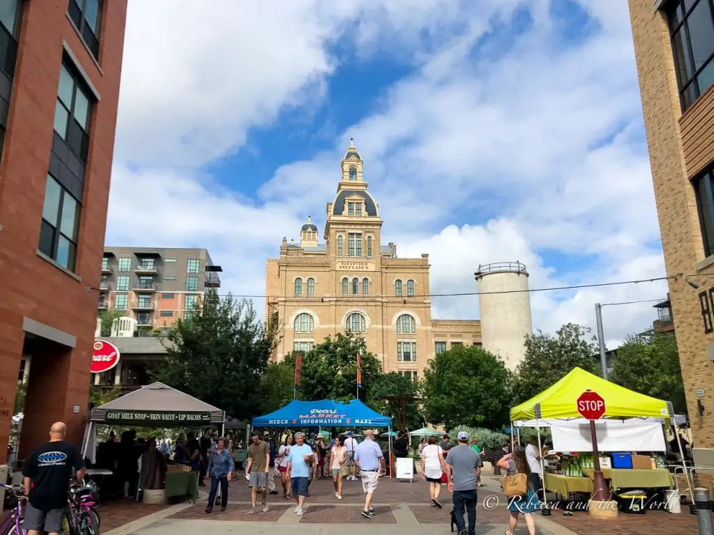 A bustling scene at the Pearl District in San Antonio. People are walking around an open area lined with vendor stalls, and the historic Pearl Brewery building, with its distinctive architecture, is prominent in the background under a blue sky with scattered clouds. The Pearl in San Antonio is a great place to visit on the weekend.