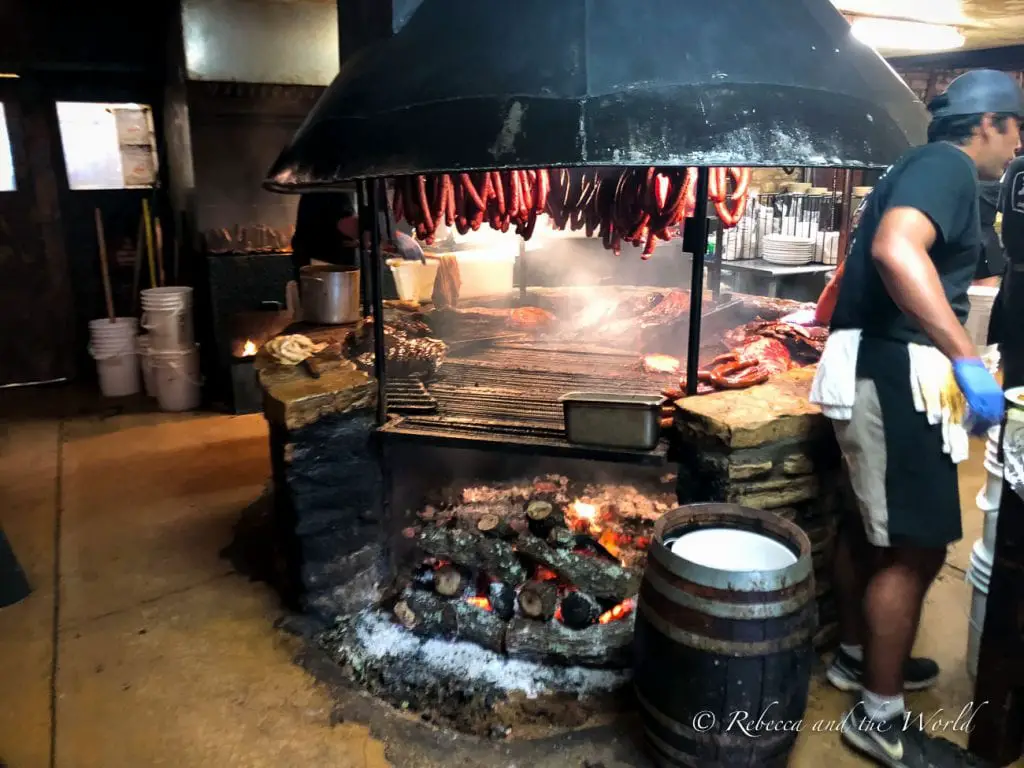 Inside a traditional Texas barbecue restaurant, showcasing a large open pit filled with smoking meats. Sausages hang from hooks above the grill, and a worker in a black shirt and white apron is tending to the food. You have to visit Salt Lick BBQ when you visit Austin!