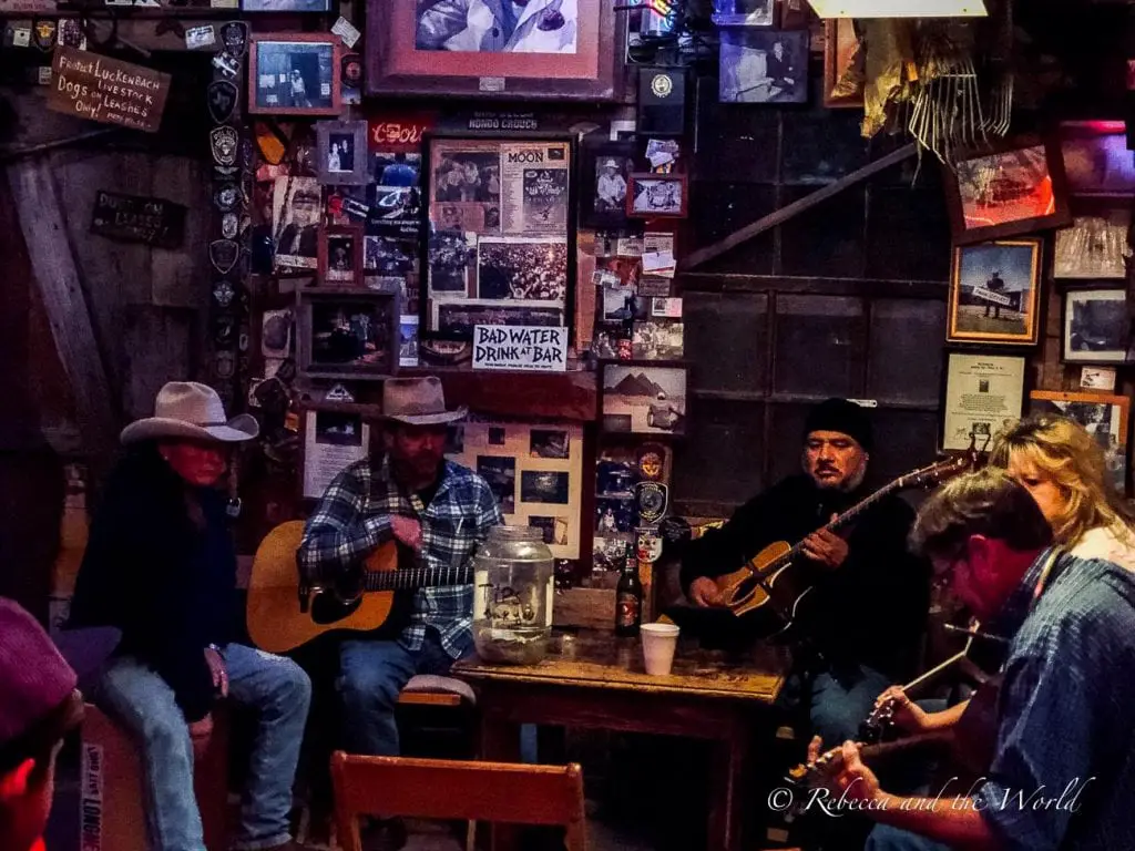 A group of musicians playing instruments in a cozy, dimly-lit room adorned with numerous photos and memorabilia on the walls. The musicians are wearing casual clothing, some with cowboy hats. Stop by Luckenbach near Fredericksburg in Texas for a fun night out.