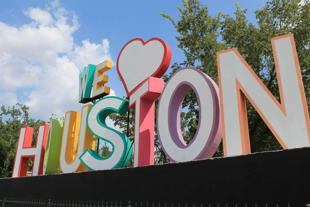 A colorful "We Love Houston" sign with large, vibrant letters. The sign is outdoors with trees and a blue sky with some clouds in the background. Houston is one of the best cities in Texas to visit.