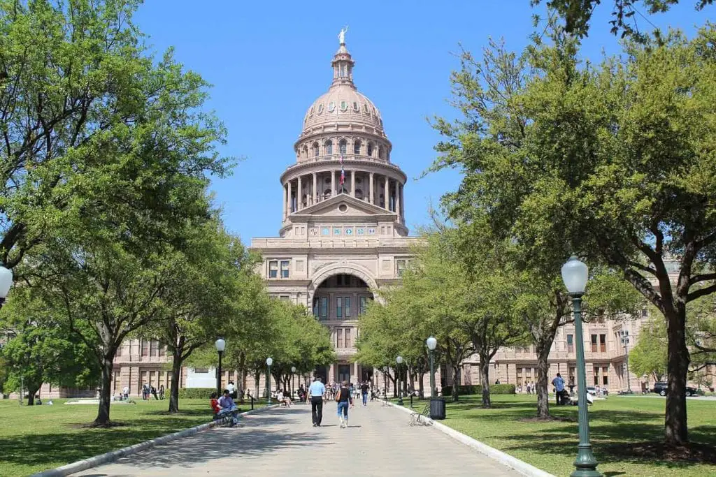The Texas State Capitol building in Austin, seen from a distance along a tree-lined walkway. The Capitol's large dome is the focal point, surrounded by greenery and a bright blue sky. Austin's Capitol building is actually taller than the capitol in Washington D.C.