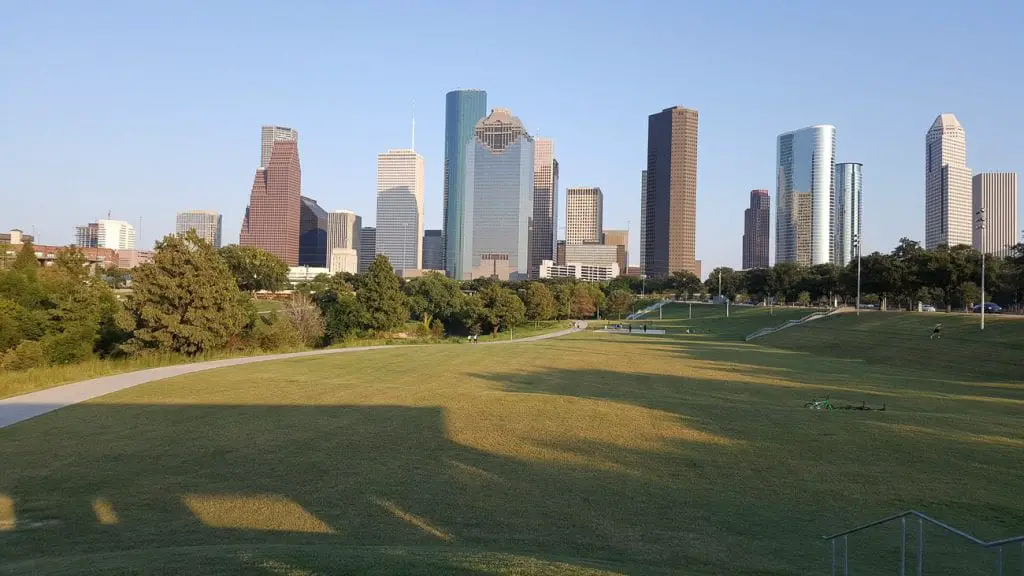 The Houston skyline during the day, featuring modern skyscrapers of various shapes and sizes. The foreground includes a grassy park area with trees and a walking path. Houston has a surprising number of green spaces.