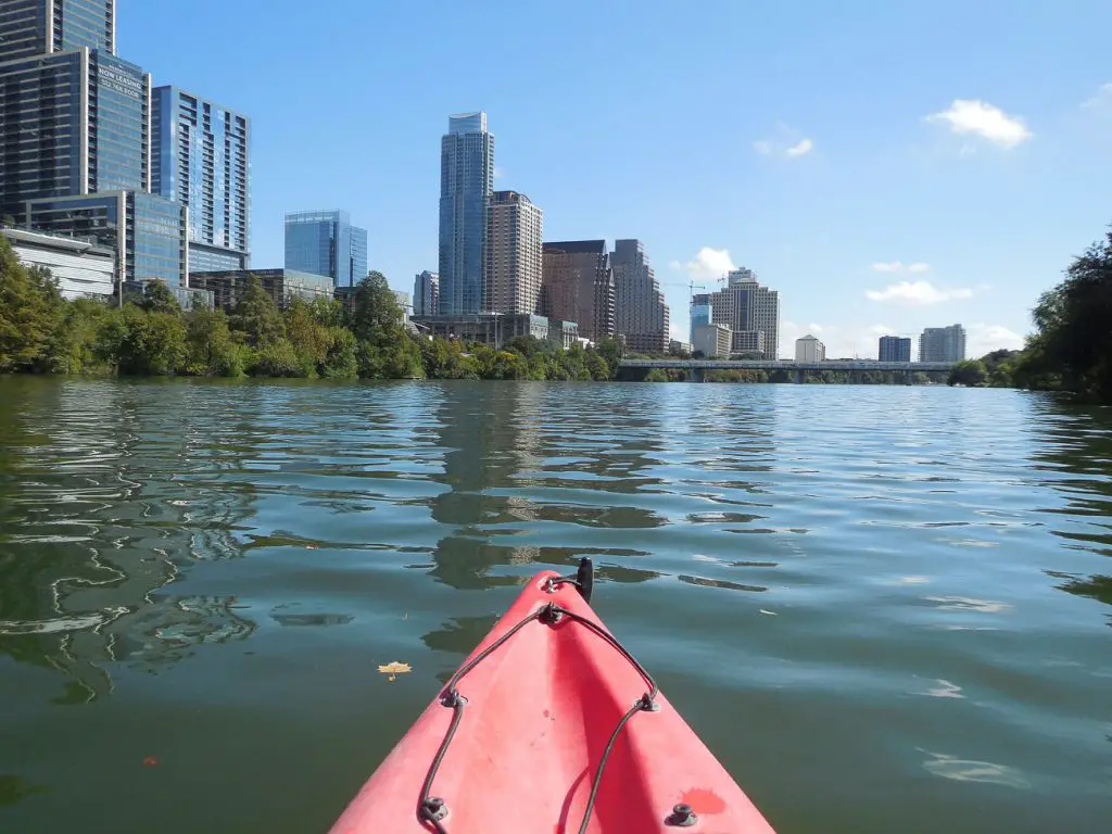 A view of the Austin skyline from Lady Bird Lake. The photo is taken from a kayak, with the red tip of the kayak visible in the foreground. Modern buildings and lush green trees line the lake's edge under a clear blue sky. Kayaking is one way to see Austin, one of the best cities in Texas.