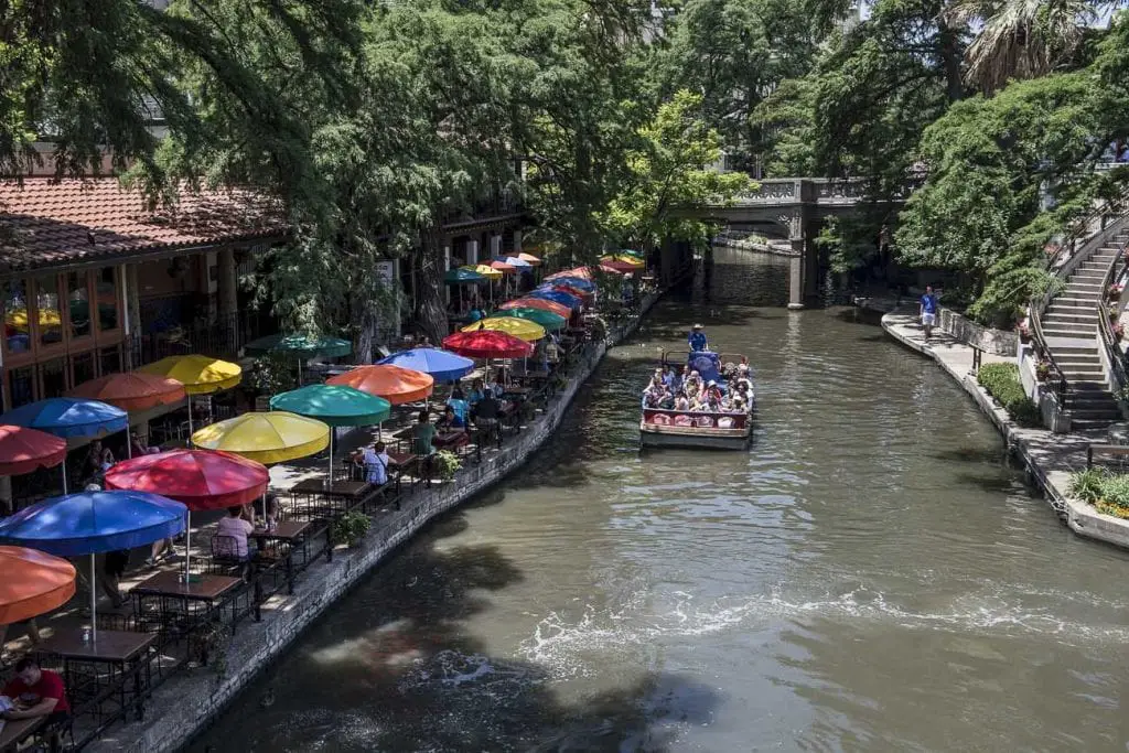 A scenic view of the San Antonio River Walk featuring a canal lined with outdoor dining tables shaded by colorful umbrellas. A tour boat filled with passengers is seen cruising down the river, and lush green trees and a stone bridge are visible in the background. San Antonio's River Walk is a must visit when in the city.