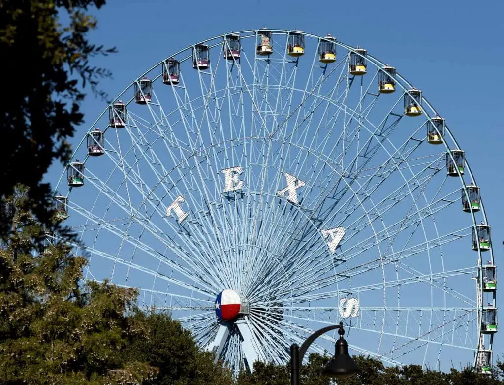 A giant ferris wheel is silhoutted against a cloudless blue sky, with TEXAS written in the middle of the wheel