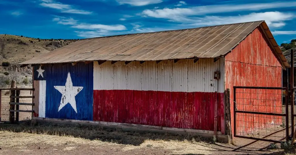 A rustic barn painted with the Texas state flag. The barn has a weathered tin roof and is situated in a rural landscape with dry grass and hills in the background under a blue sky with scattered clouds.