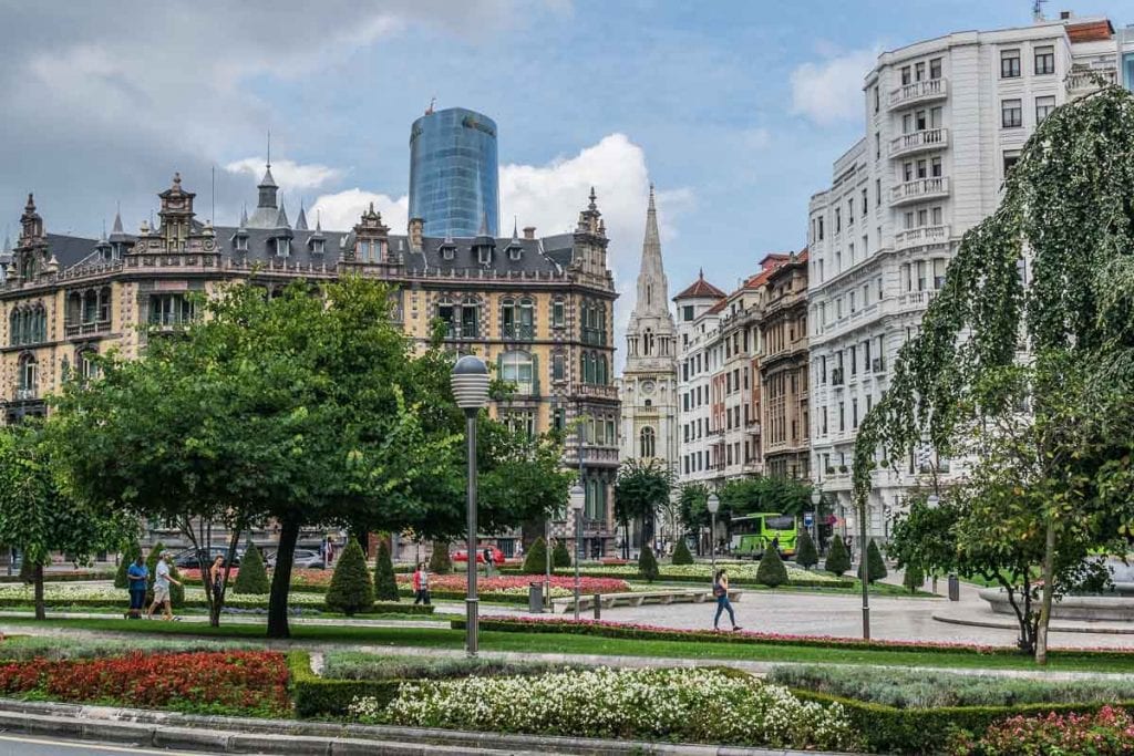 An urban park in Bilbao with manicured flowerbeds and a mix of traditional and modern architecture in the background, depicting the city's blend of green spaces and urban development.