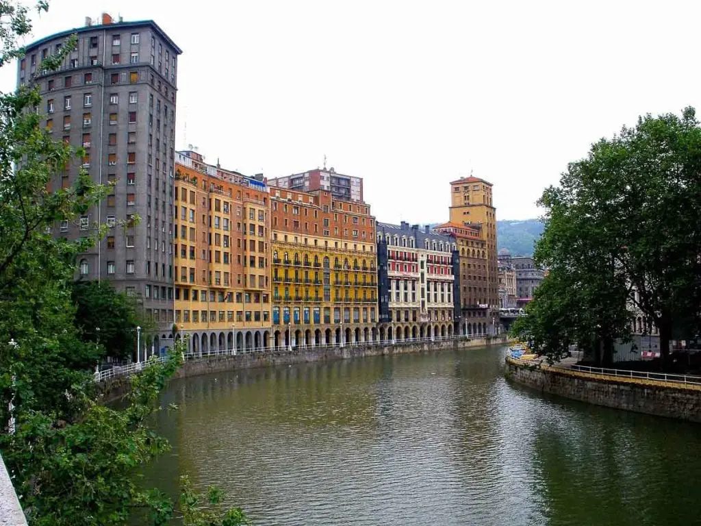 The Nervión River in Bilbao flanked by colorful apartment buildings with classical facades, reflecting a vibrant urban life alongside the tranquil water. Bilbao, Spain, sits on the Nervion River, and the architecture in the city is lovely to explore.