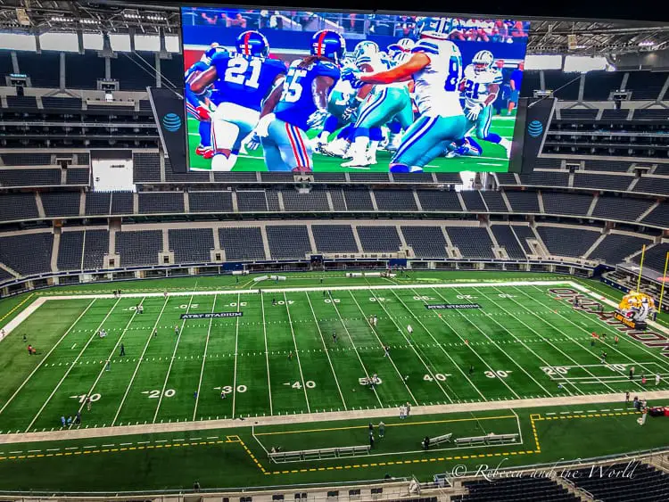 Interior view of the AT&T Stadium in Arlington, Texas, near Dallas, showing the expansive football field and a giant screen displaying a game.