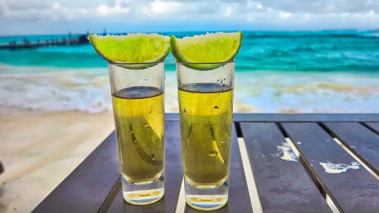 Two shot glasses with golden liquid and lime wedges on the rim, sitting on a wooden table with a soft-focus beach background.