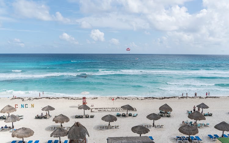 Aerial view of Cancun beach with clear turquoise waters, white sands, and thatched umbrellas providing shade to beachgoers.