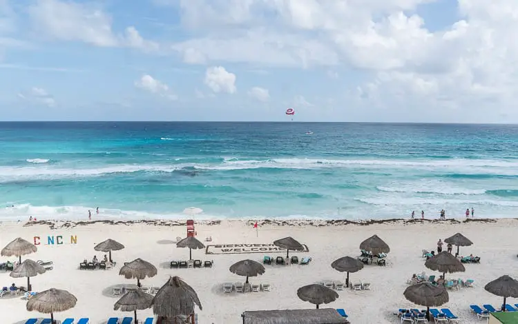 Aerial view of Cancun beach with clear turquoise waters, white sands, and thatched umbrellas providing shade to beachgoers.