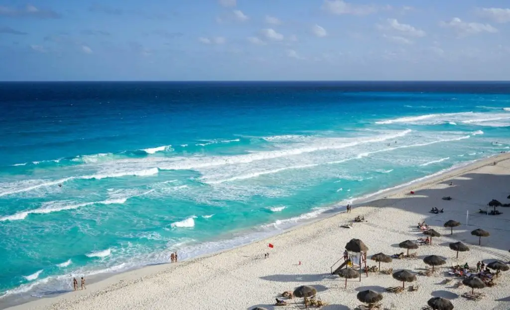 A wide angle view of a pristine Cancun beach with bright blue waters and waves gently crashing onto the sandy shore.
