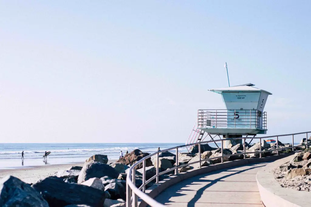 Lifeguard tower on a sunny beach in San Diego with clear skies. Two surfers and cyclists are visible in the distance on the sand. San Diego is a photographer's dream location - the beaches are beautiful and quintessentially Californian.