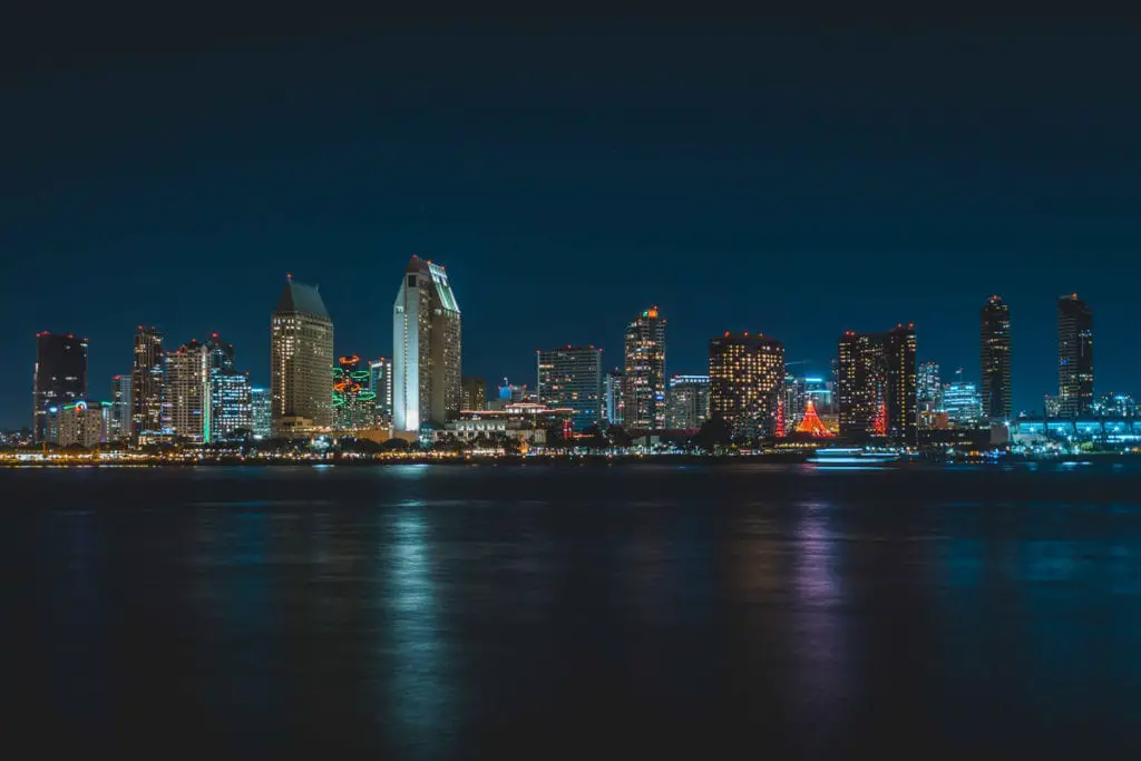 Night view of a brightly lit city skyline reflecting on a body of water, with various sizes of buildings and festive lights. The San Diego skyline is a sight to behold.