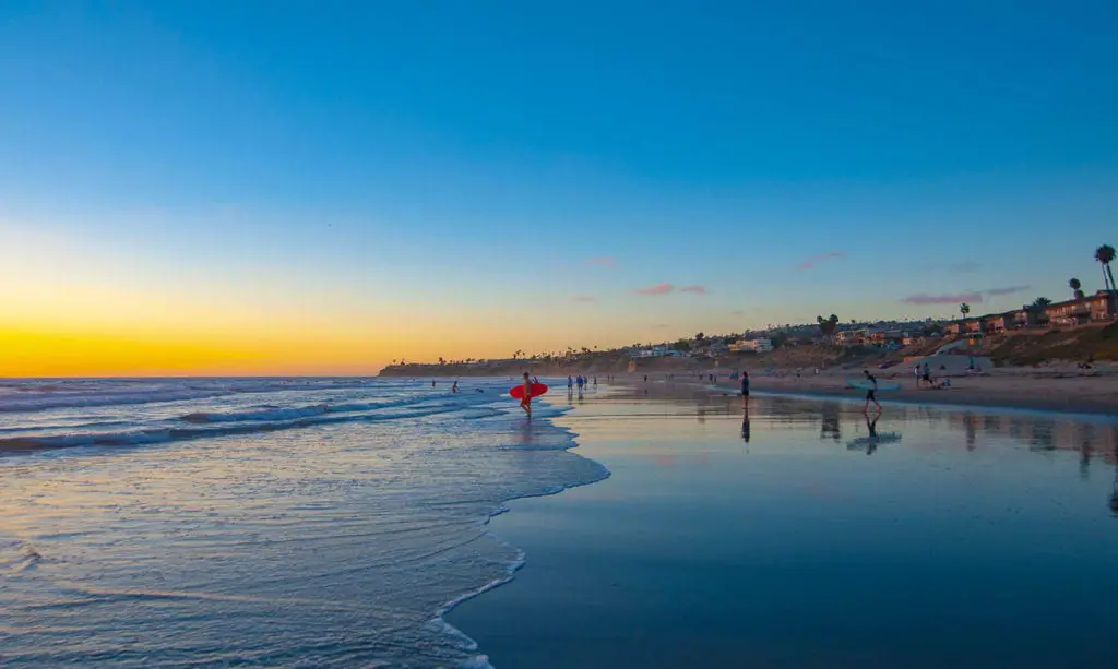 Beach scene at sunset with people walking along the shore, surfers in the water, and a vibrant sky reflecting on the wet sand. There are so many beaches to add to your San Diego itinerary, you could easily spend a few days or a week exploring them all.