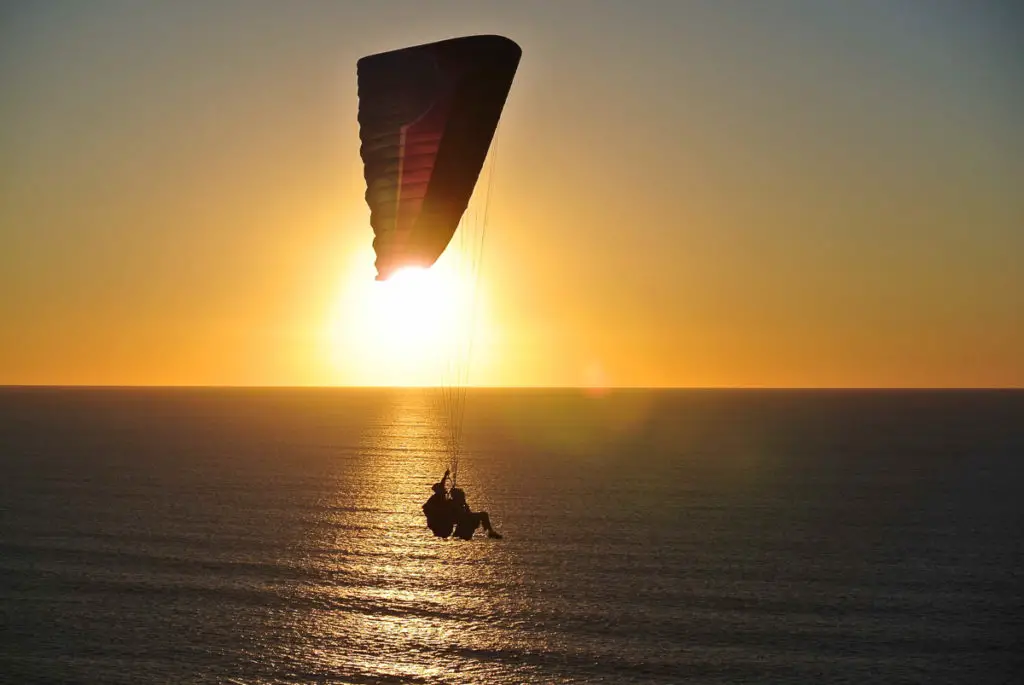 A paraglider in silhouette against a sunset sky, over a calm sea, with the sun reflecting on the water's surface. The are plenty of great beachy things to do in San Diego, including paragliding.