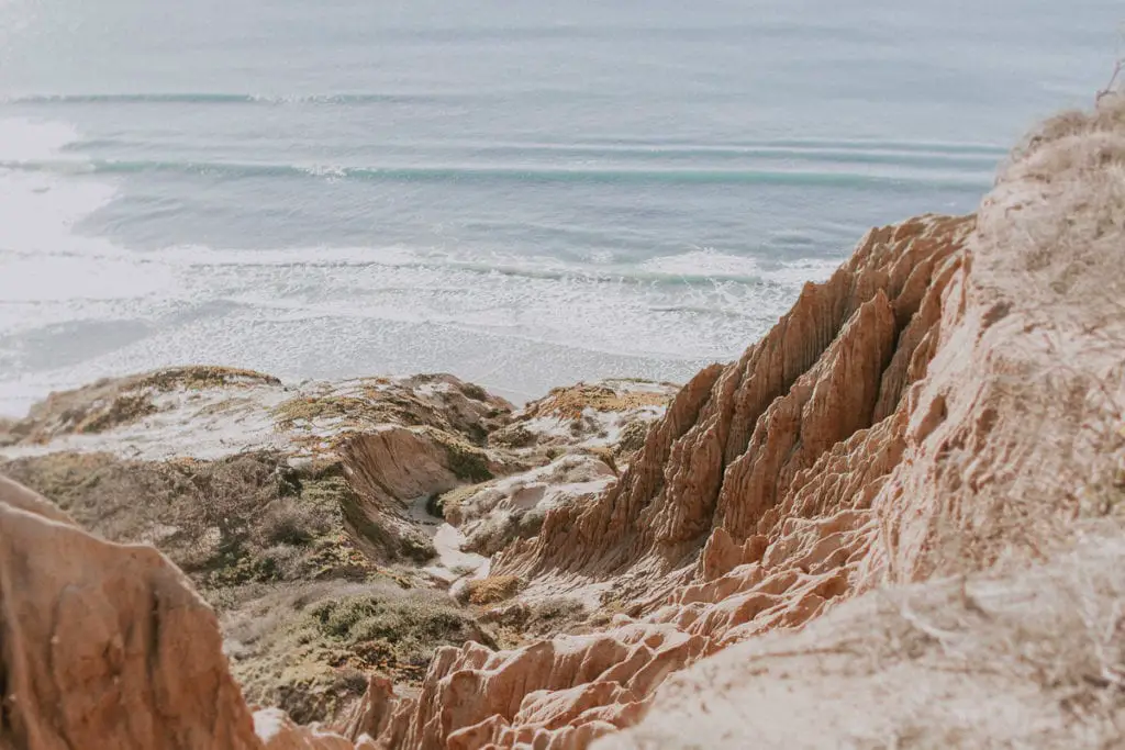 View from atop a cliff overlooking a beach with gentle waves. The foreground shows textured, eroded cliffs with sparse vegetation. Torrey Pines in San Diego is a great place for hikes with epic views.