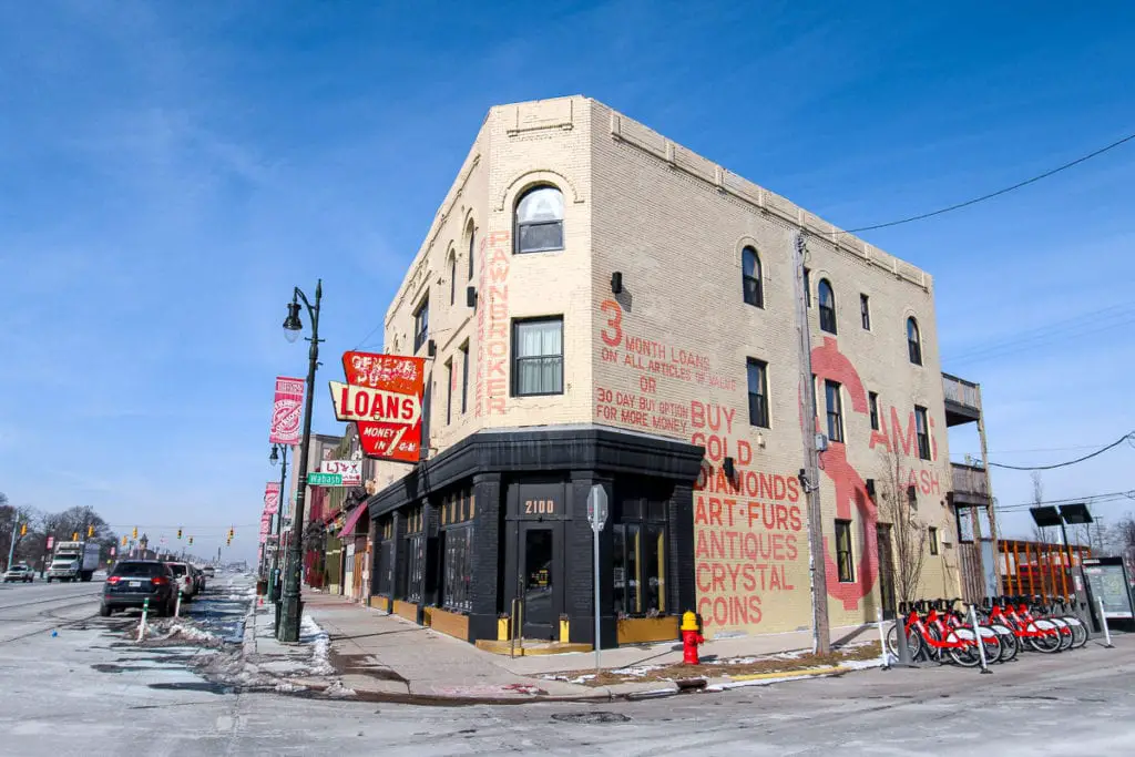 Street corner in Detroit with a two-story building featuring vibrant pawnbroker signage, against a backdrop of a clear blue sky. One of the fun things to do in Detroit is have a drink at one of the hole-in-the-wall places around the city.