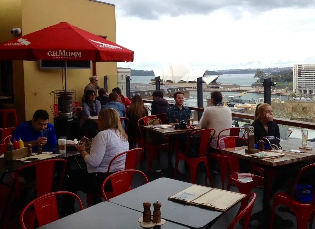 Casual rooftop dining scene with patrons seated at tables, red umbrellas, and a view of the Sydney Opera House and harbour in the background, indicative of Sydney's vibrant cafe culture. Make sure to get a pub meal when you visit Sydney.