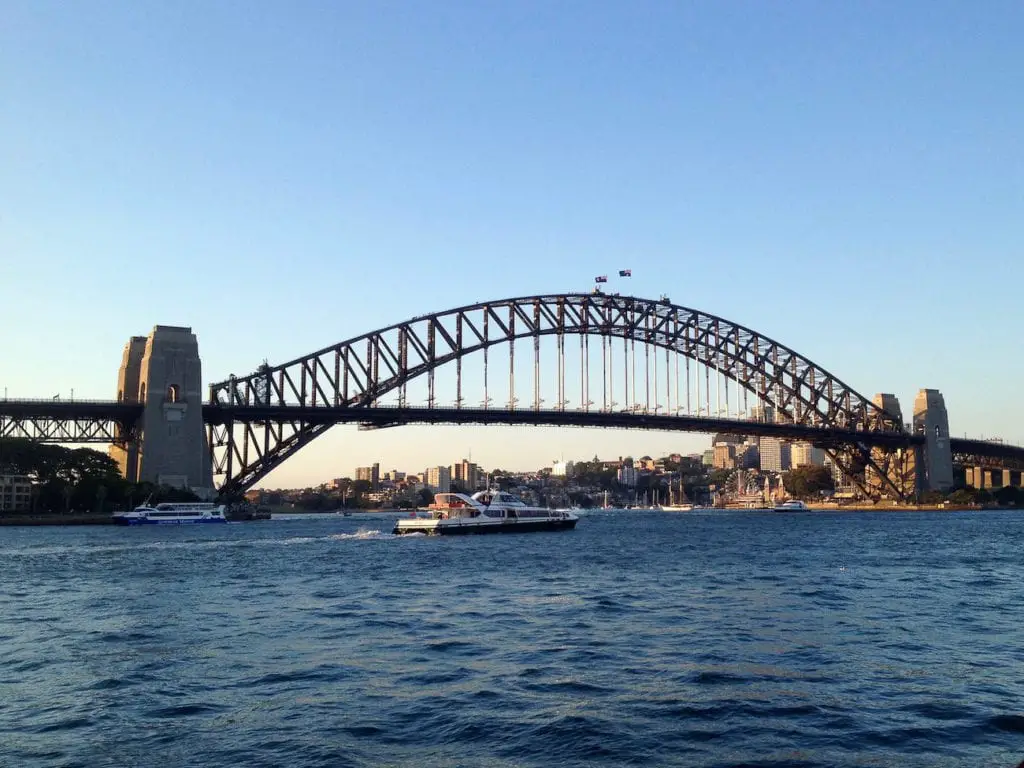 Panoramic daytime view of the Sydney Harbour Bridge from a waterfront perspective, with the Australian flag atop, against a clear sky and the Sydney skyline in the distance. One of the most iconic sights in Sydney is the Sydney Harbour Bridge.