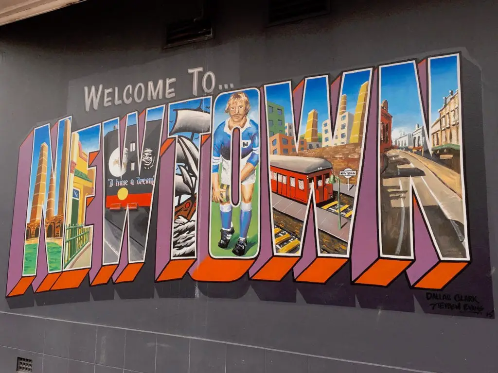 Colourful street mural reading 'Welcome To... Newtown' featuring various scenes of the suburb's landmarks and culture, reflecting the area's artistic and eclectic vibe. Newtown in Sydney has some cool street art to explore.