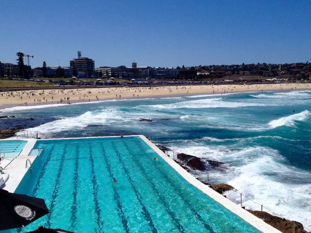 Bondi Icebergs ocean pool with turquoise waters, bordering the crashing waves of the sea, while sunbathers and swimmers enjoy the sandy Bondi Beach in the background. A trip to Bondi Beach is one of the top things to do in Sydney.