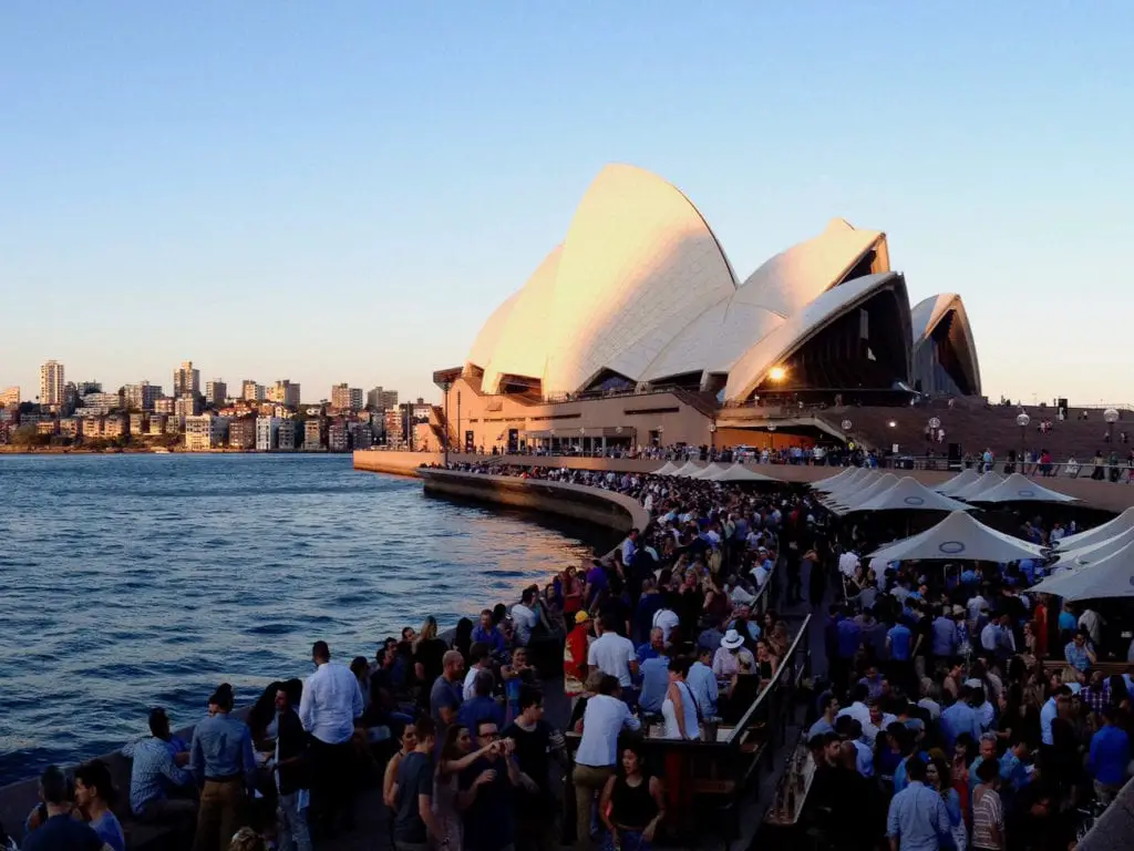 Sydney Opera House at sunset, its distinctive sail-like design illuminated, with crowds gathered on the steps and waterfront area, as seen from the bustling Circular Quay. Every Sydney itinerary should include a visit to the Sydney Opera House.