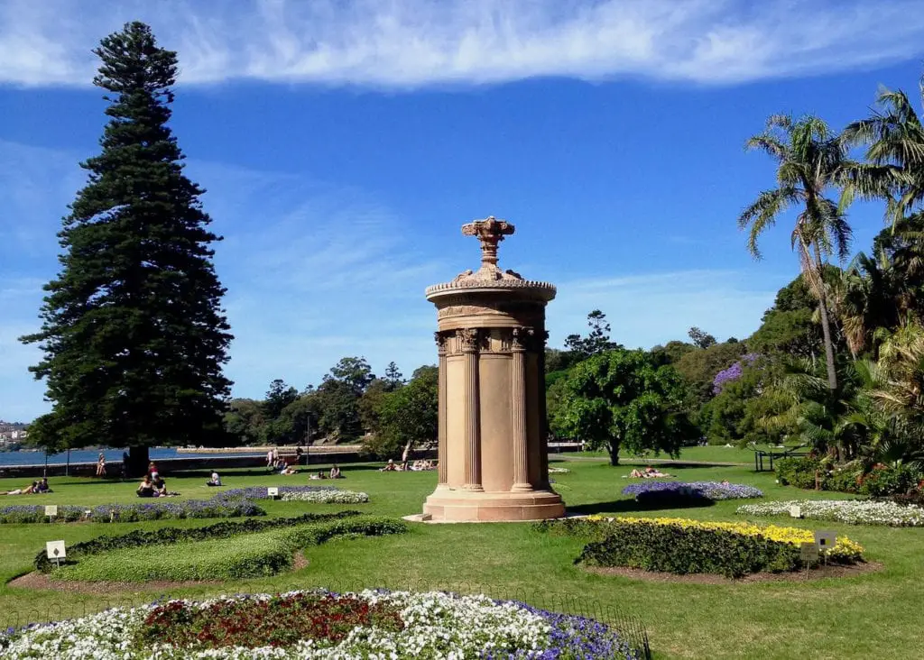 Lush greenery of the Royal Botanic Garden with a large, ornate sandstone Choragic Monument, surrounded by manicured flower beds and visitors enjoying the scenery. Sydney's Royal Botanic Gardens are a must-visit, especially to cool down on a hot day.