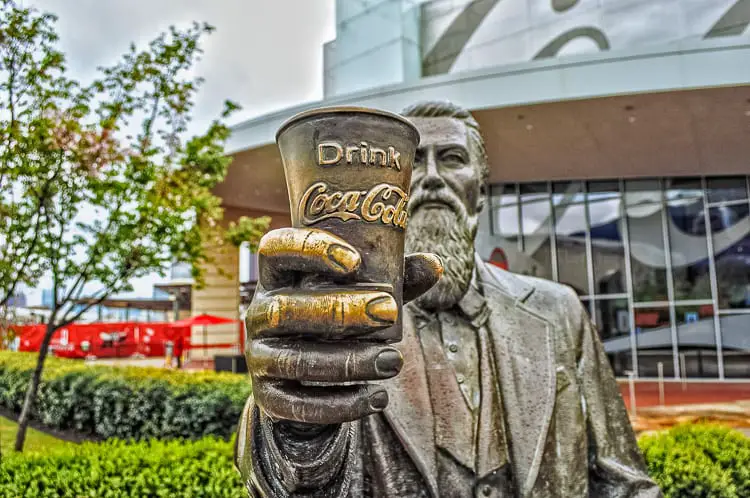 Statue of a bearded man holding out a weathered Coca-Cola cup, with trees and the facade of a modern building in the background.