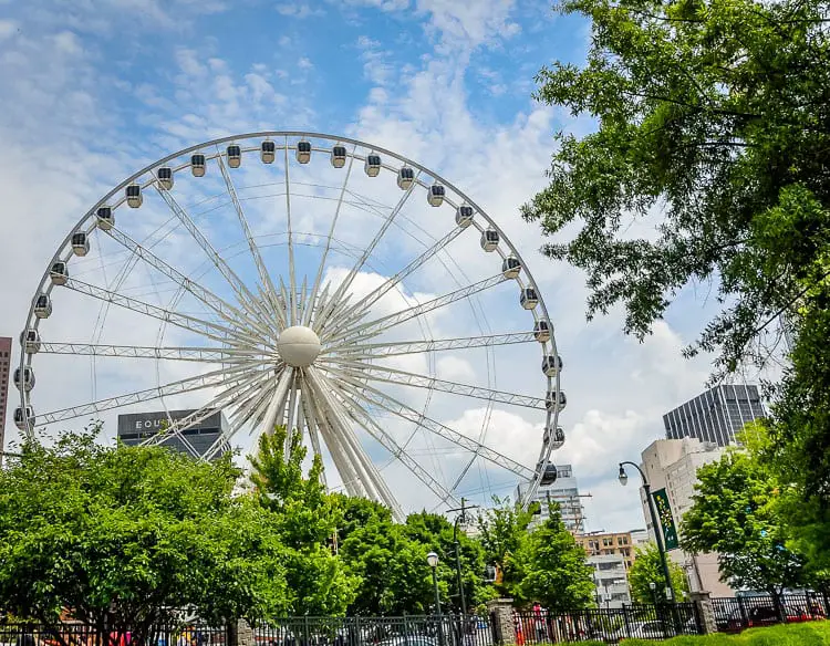 A large Ferris wheel with enclosed gondolas set against a partly cloudy sky, surrounded by green foliage and urban buildings. The Skyview Atlanta is a great thing to do on a weekend in Atlanta for amazing views of the city.